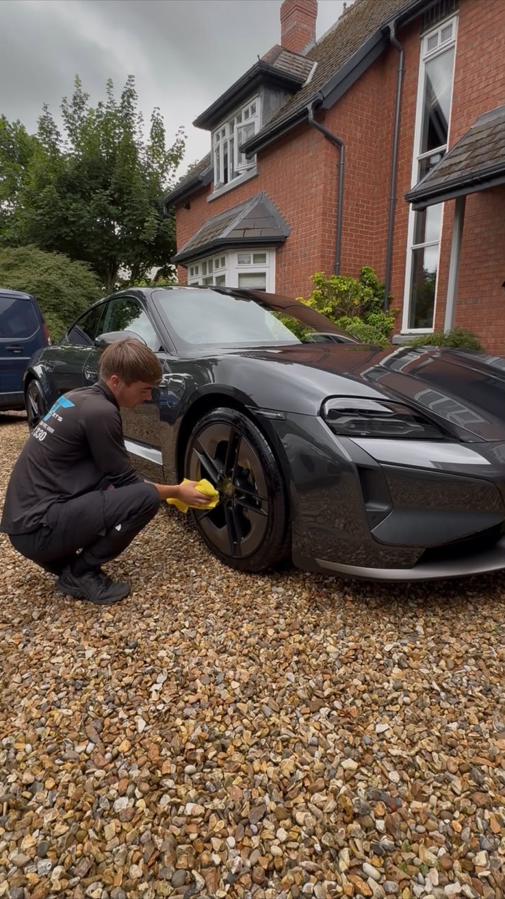 ST Valets detailer cleaning the alloy wheel of a Porsche Taycan on a gravel driveway